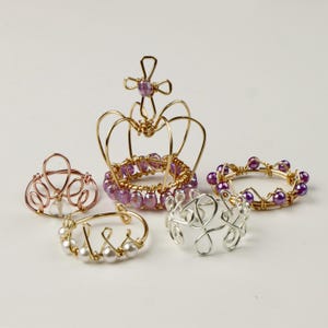 May include: Five wire rings with different designs. The largest ring is a gold crown with purple beads and a cross on top. The other rings are smaller and have different designs, including a heart shape and a circle with beads.