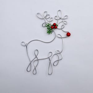 May include: A silver wire sculpture shaped like a reindeer silhouette. The design features looped wire antlers, a red bead nose, and a cluster of green wire leaves with three small red beads representing holly berries attached to the antlers.