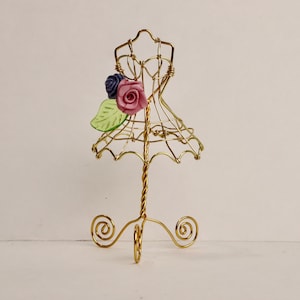 May include: A gold wire dress form with a pink and purple flower accent. The dress form is on a gold wire stand with three legs.