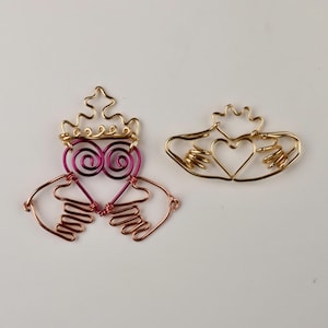 Op de afbeelding: Twee draadbeeldjes van de Claddagh, een traditioneel Iers symbool. Eén is goud met een hart en handen, de andere is goud met een roze hart en handen.