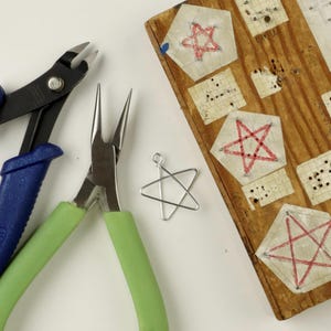 May include: Two pairs of pliers, one with blue handles and one with green handles, are used to create a wire star. The star is laying on a white surface. A wooden board with red star patterns is in the background.