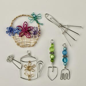 Peut inclure: Un ensemble de cinq outils de jardin miniatures en fil de fer, comprenant un arrosoir, une pelle, un râteau, une fourche et un panier de fleurs. Les outils sont en fil d'argent et le panier est en fil d'or. Les fleurs dans le panier sont violettes, roses et bleues.