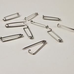 May include: A group of ten silver safety pins scattered on a white surface. The pins are all the same size and shape, with a simple design.