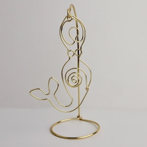 May include: Gold wire sculpture of a mermaid on a stand. The mermaid is depicted in a stylized, abstract form with a flowing tail and a circular design in the center of her body.