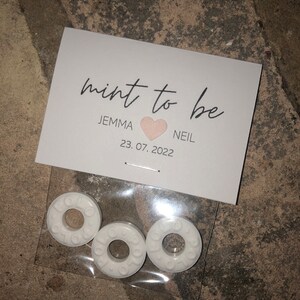 May include: White favor bags with the text "mint to be" and a pink heart. The bags contain three white round mints. The text below the heart reads "Jemma & Neil 23.07.2022".