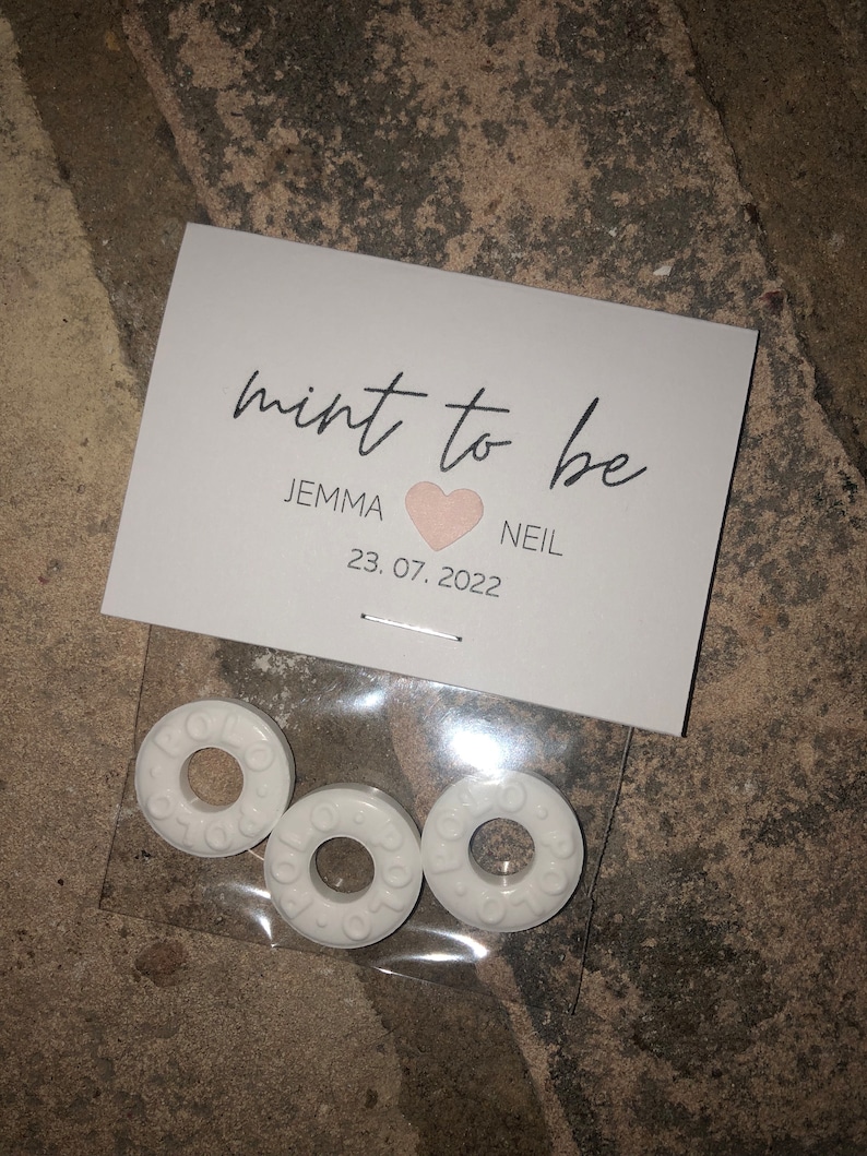 May include: White favor bags with the text "mint to be" and a pink heart. The bags contain three white round mints. The text below the heart reads "Jemma & Neil 23.07.2022".