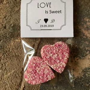May include: Two pink heart-shaped chocolates with white sprinkles, packaged in a clear plastic bag with a white card that reads "LOVE Is Sweet" and "I ♥ D 23.09.2019".