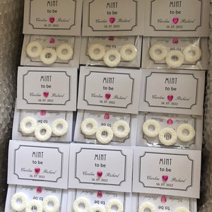 May include: Nine white favor boxes with clear cellophane windows, each containing three round white mints with the text "Mint to be" and a pink heart with a rhinestone. The boxes also include a white card with the text "Mint to be" and the names "Caroline" and "Richard" with the date "16.07.2022".