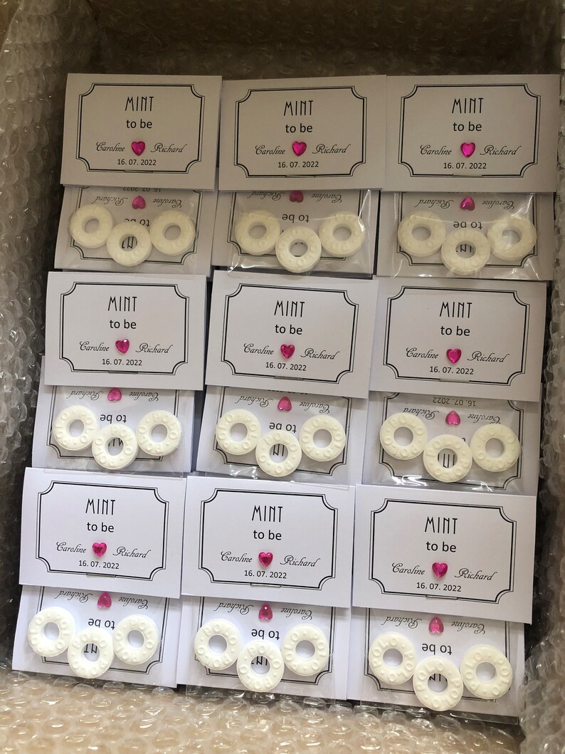 May include: Nine white favor boxes with clear cellophane windows, each containing three round white mints with the text "Mint to be" and a pink heart with a rhinestone. The boxes also include a white card with the text "Mint to be" and the names "Caroline" and "Richard" with the date "16.07.2022".