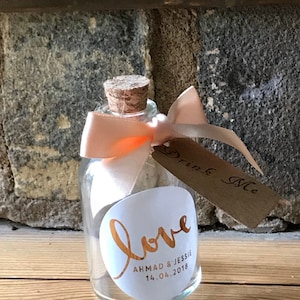 May include: A small glass bottle with a cork stopper and a peach-colored ribbon tied around the neck. The bottle has a white label with the word "love" written in gold script and the text "Ahmad & Jessie 14.04.2018". A brown tag with the text "Drink Me" is attached to the ribbon.