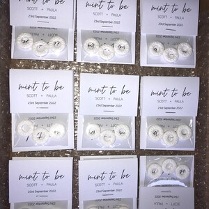 May include: Nine white favor boxes with clear cellophane windows. Each box contains three white round mints with the text "mint to be" and "SCOTT + PAULA" and "23rd September 2022" printed on the front.