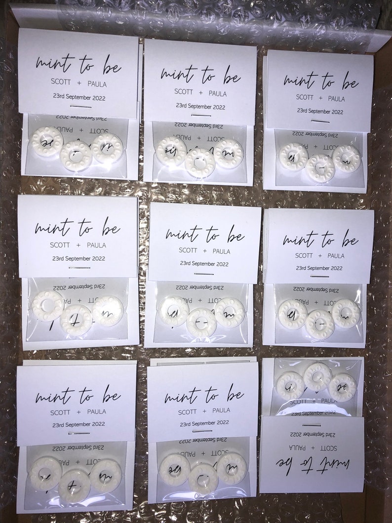May include: Nine white favor boxes with clear cellophane windows. Each box contains three white round mints with the text "mint to be" and "SCOTT + PAULA" and "23rd September 2022" printed on the front.