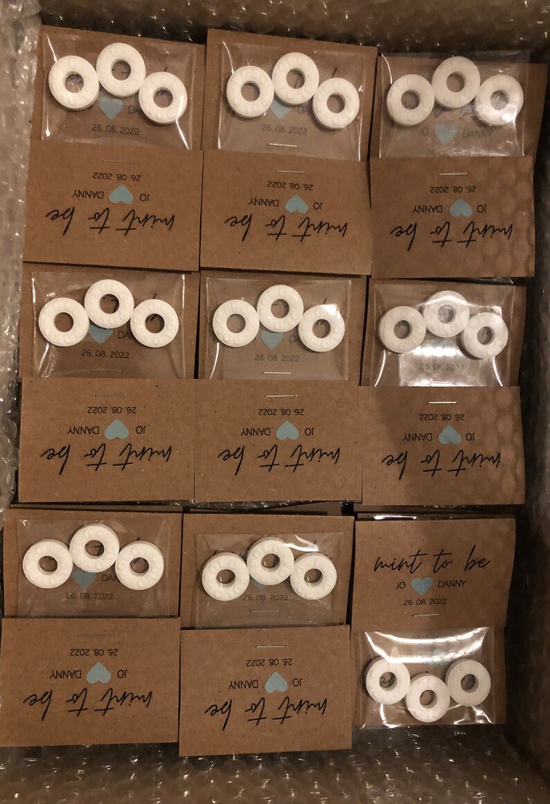 May include: A set of small, white, round mints with a hole in the center, packaged in clear plastic bags with brown paper cards. The cards have the text "mint to be" with the date and names "JO & DANNY" printed on them.
