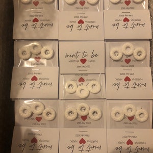 May include: Mint to be wedding favors with white round mints and a white card with the text "mint to be" in black cursive writing, "MARTINA" and "MARK" in black text, and "29th July 2022" in black text.