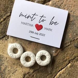 May include: A white card with the text "mint to be" in black cursive writing. Below the text, it says "Martina" followed by a red heart and "Mark". The date "29th July 2022" is printed below the names. Three white round mints are in a clear plastic bag.