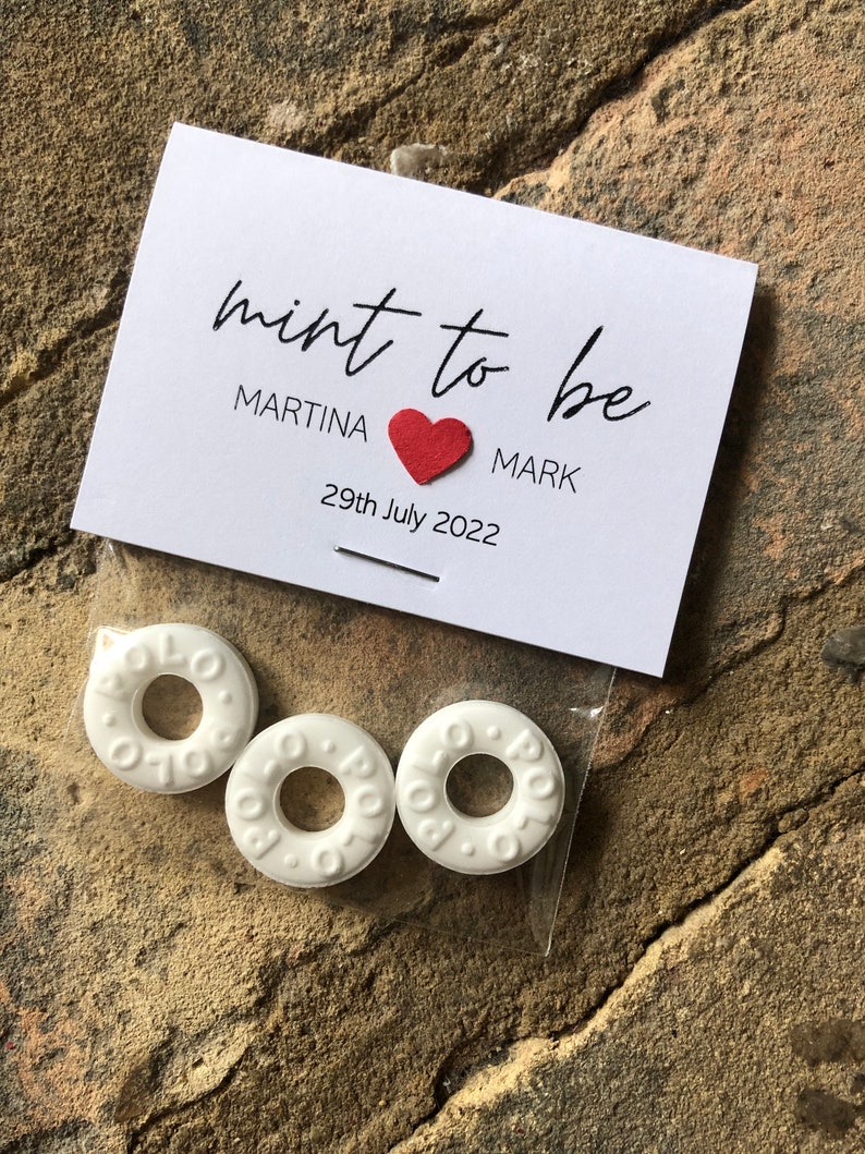 May include: A white card with the text "mint to be" in black cursive writing. Below the text, it says "Martina" followed by a red heart and "Mark". The date "29th July 2022" is printed below the names. Three white round mints are in a clear plastic bag.