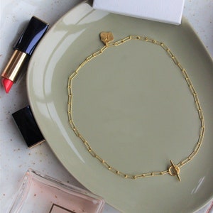 Può includere: Una collana a catena d'oro con un piccolo ciondolo d'oro e una chiusura a levetta. La collana è su un piatto verde chiaro. Il piatto è su una superficie bianca.