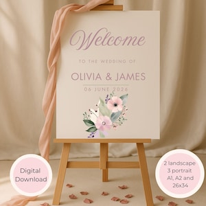May include: A wedding welcome sign on a wooden easel. The sign is cream-colored with the word "Welcome" in a soft purple script. Below, it reads "TO THE WEDDING OF OLIVIA & JAMES 06 JUNE 2026." A floral arrangement decorates the bottom. A pink ribbon drapes from the top.
