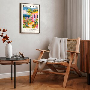 May include: A framed poster with a colorful illustration of a pink house with a palm tree and ocean view. The poster is titled "Henri Matisse" and has the text "Berggruen & Cie" at the bottom. The poster is hanging on a white wall above a wooden chair with a white throw blanket.