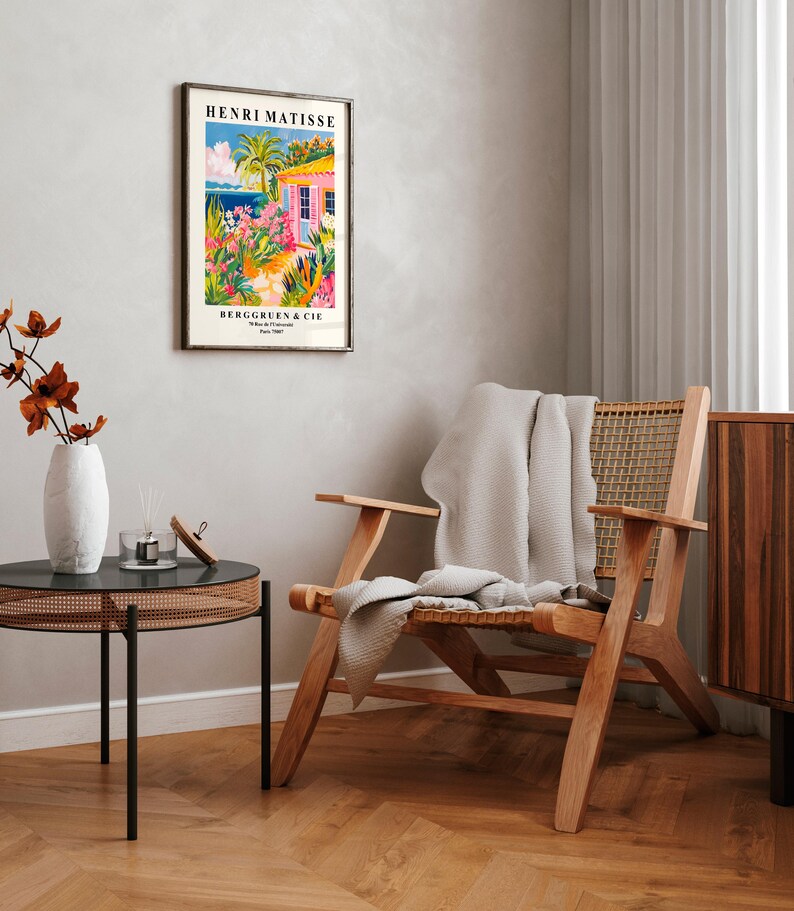 May include: A framed poster with a colorful illustration of a pink house with a palm tree and ocean view. The poster is titled "Henri Matisse" and has the text "Berggruen & Cie" at the bottom. The poster is hanging on a white wall above a wooden chair with a white throw blanket.