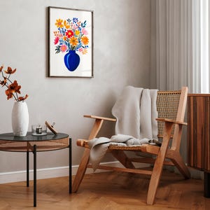 Op de afbeelding: Een ingelijst aquarel schilderij van een boeket kleurrijke bloemen in een blauwe vaas. Het schilderij hangt aan een witte muur in een woonkamer met een houten stoel en een bijzettafel.