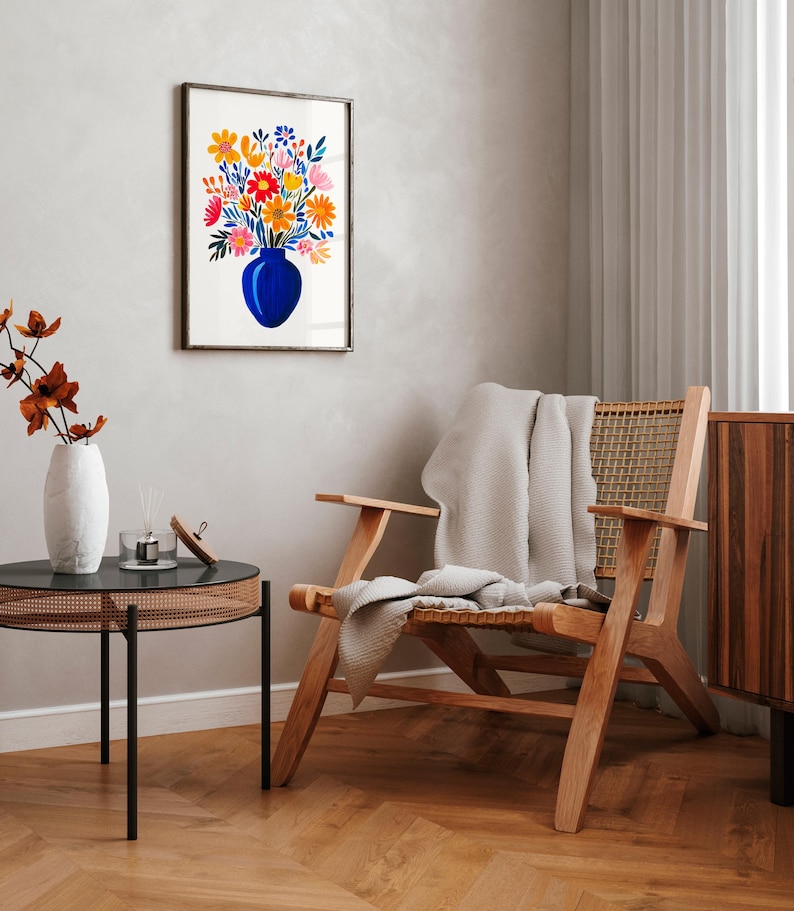 Op de afbeelding: Een ingelijst aquarel schilderij van een boeket kleurrijke bloemen in een blauwe vaas. Het schilderij hangt aan een witte muur in een woonkamer met een houten stoel en een bijzettafel.