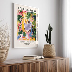 May include: A framed Henri Matisse art print featuring a colorful illustration of a person reading a newspaper in a tropical setting. The print includes the text "HENRI MATISSE" and "BERGGRUEN & CIE". A cactus in a woven basket sits on a wooden cabinet.