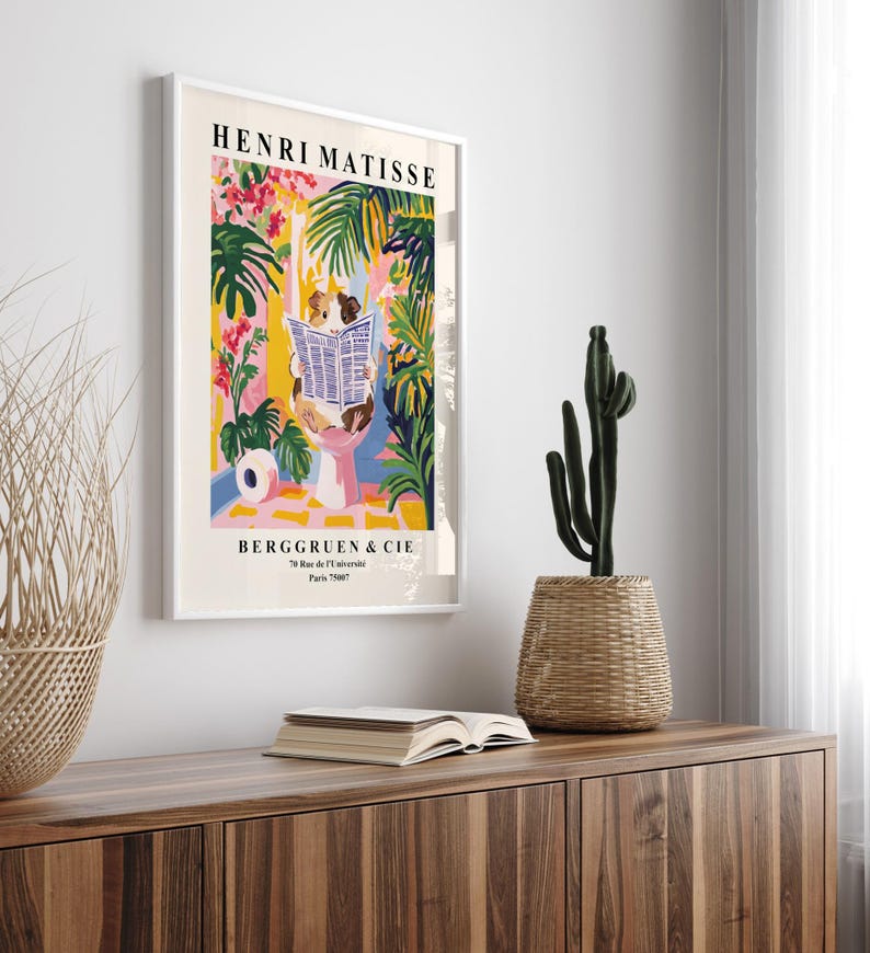 May include: A framed Henri Matisse art print featuring a colorful illustration of a person reading a newspaper in a tropical setting. The print includes the text "HENRI MATISSE" and "BERGGRUEN & CIE". A cactus in a woven basket sits on a wooden cabinet.