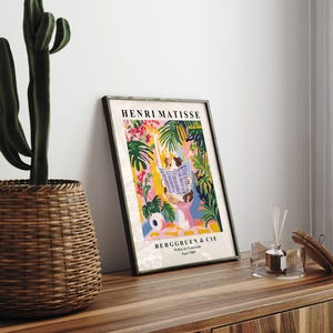 May include: Framed Henri Matisse art print featuring a colorful illustration of a person reading a newspaper amidst tropical foliage. The print includes the text "BERGGRUEN & CIE" and is displayed on a wooden surface.
