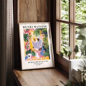 May include: Framed art print featuring a colorful illustration with the text "HENRI MATISSE" and "BERGGRUEN & CIE". The artwork depicts a person reading a newspaper amidst vibrant tropical plants in shades of pink, green, and yellow. The print is displayed on a wooden surface.