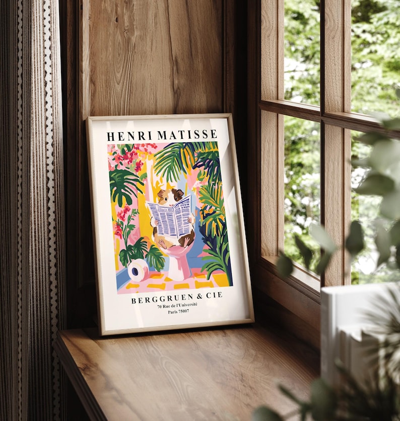 May include: Framed art print featuring a colorful illustration with the text "HENRI MATISSE" and "BERGGRUEN & CIE". The artwork depicts a person reading a newspaper amidst vibrant tropical plants in shades of pink, green, and yellow. The print is displayed on a wooden surface.