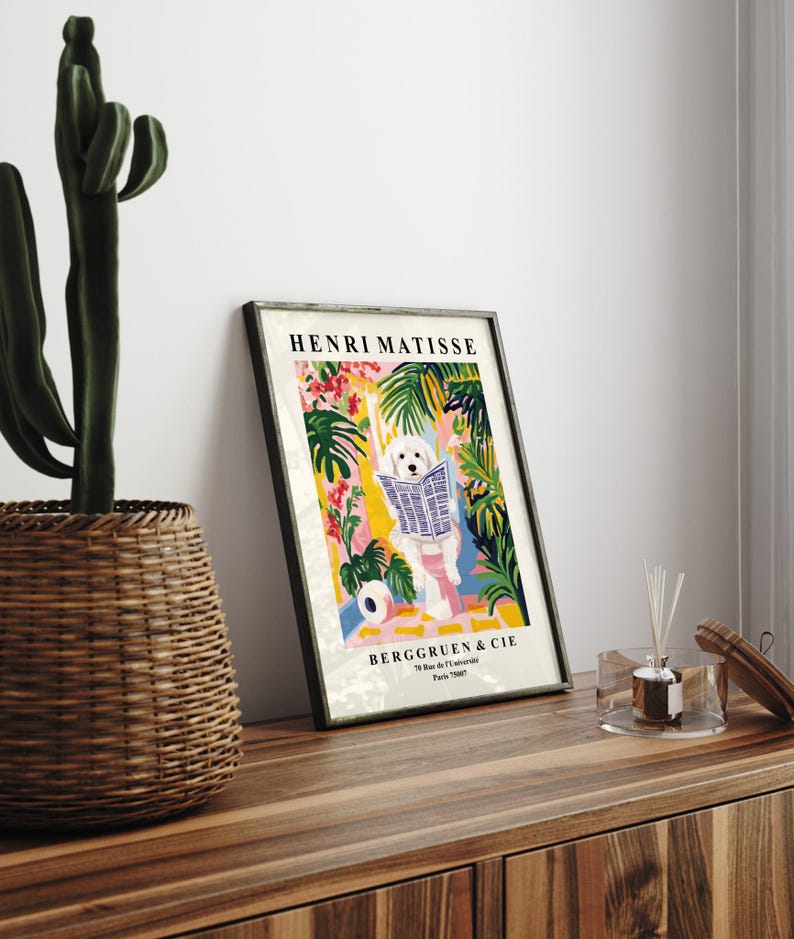 May include: A framed art print depicting a white dog reading a newspaper amidst a vibrant tropical scene. The print is labelled "HENRI MATISSE" and includes the text "BERGGRUEN & CIE". The artwork is displayed on a wooden surface, with a cactus in a basket.