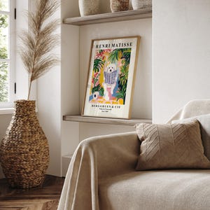 May include: A framed Henri Matisse art print showing a white dog reading a newspaper, set against a colourful tropical background. The print is displayed on a shelf above a sofa with a decorative pillow. A woven vase with dried plants is in the foreground.