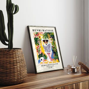 May include: Framed art print featuring a dog reading a newspaper, surrounded by colorful tropical foliage. The print is titled "HENRI MATISSE" and includes the text "BERGGRUEN & CIE". The artwork is displayed on a wooden surface.