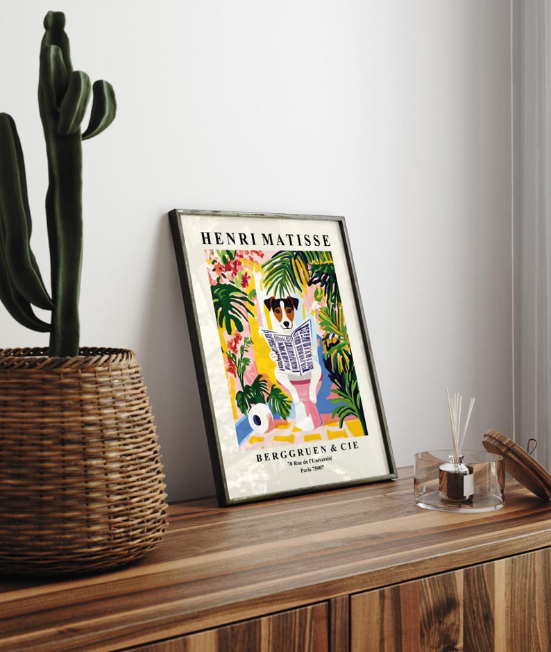 May include: Framed art print featuring a dog reading a newspaper, surrounded by colorful tropical foliage. The print is titled "HENRI MATISSE" and includes the text "BERGGRUEN & CIE". The artwork is displayed on a wooden surface.