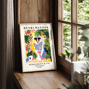 May include: A framed art print featuring a dog reading a newspaper, set against a colorful backdrop of plants and flowers. The print includes the text "HENRI MATISSE" and "BERGGRUEN & CIE". The artwork is displayed on a wooden surface near a window.