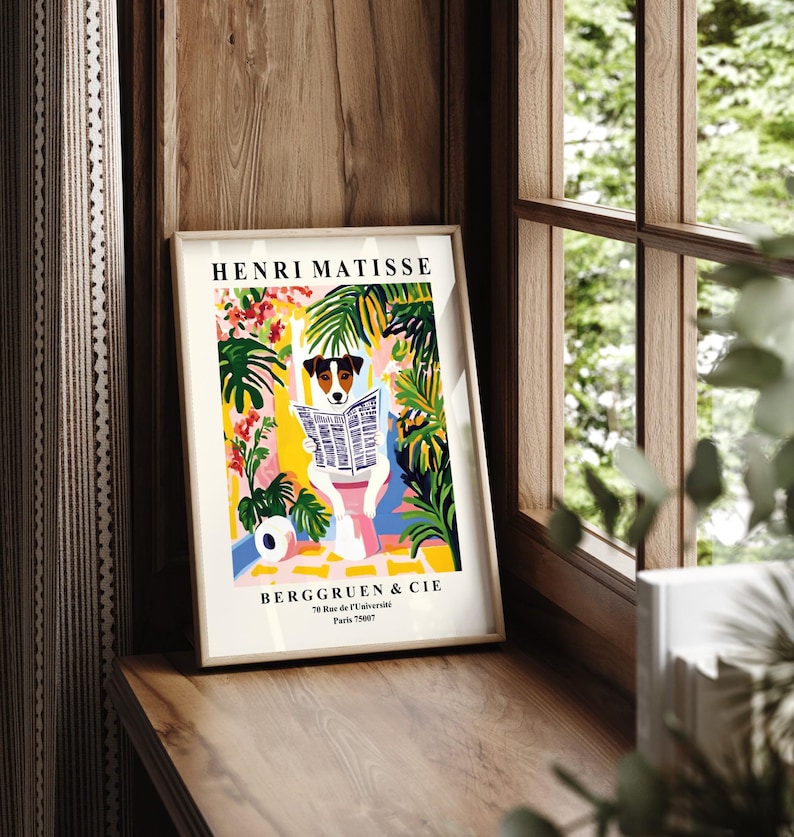 May include: A framed art print featuring a dog reading a newspaper, set against a colorful backdrop of plants and flowers. The print includes the text "HENRI MATISSE" and "BERGGRUEN & CIE". The artwork is displayed on a wooden surface near a window.
