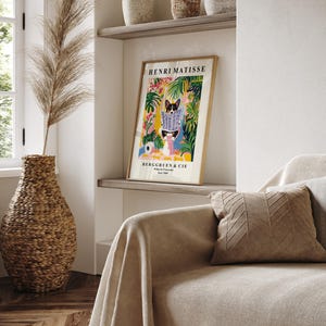 Puede incluir: Un cuadro enmarcado de Henri Matisse con un perro leyendo un peri&oacute;dico, sobre un fondo floral colorido. El cuadro se exhibe en una estanter&iacute;a sobre un sof&aacute; con un coj&iacute;n decorativo. Un jarr&oacute;n tejido con hierba seca alta est&aacute; en primer plano.