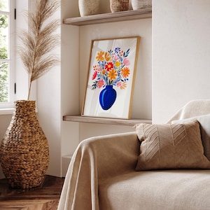 Op de afbeelding: Een ingelijst afdruk van een blauwe vaas gevuld met kleurrijke bloemen. De vaas staat op een witte achtergrond. De afdruk hangt aan een muur in een kamer met een bank en een rieten vaas.