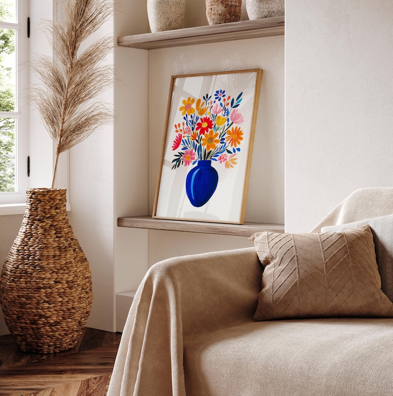 Op de afbeelding: Een ingelijst afdruk van een blauwe vaas gevuld met kleurrijke bloemen. De vaas staat op een witte achtergrond. De afdruk hangt aan een muur in een kamer met een bank en een rieten vaas.