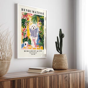 May include: A framed art print depicting a white dog reading a newspaper, set amidst vibrant plants and flowers. The print is titled "HENRI MATISSE" and includes the text "BERGGRUEN & CIE". The artwork is displayed on a wooden cabinet, with a cactus in a woven pot.