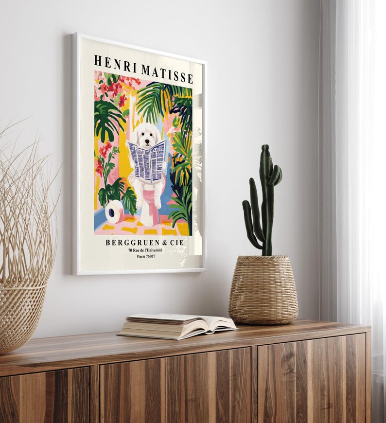 May include: A framed art print depicting a white dog reading a newspaper, set amidst vibrant plants and flowers. The print is titled "HENRI MATISSE" and includes the text "BERGGRUEN & CIE". The artwork is displayed on a wooden cabinet, with a cactus in a woven pot.