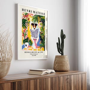 May include: A framed art print featuring a dog reading a newspaper, with the text "HENRI MATISSE" at the top. The artwork has a colorful, floral background. The print is displayed on a wooden cabinet, with a cactus and a woven basket.