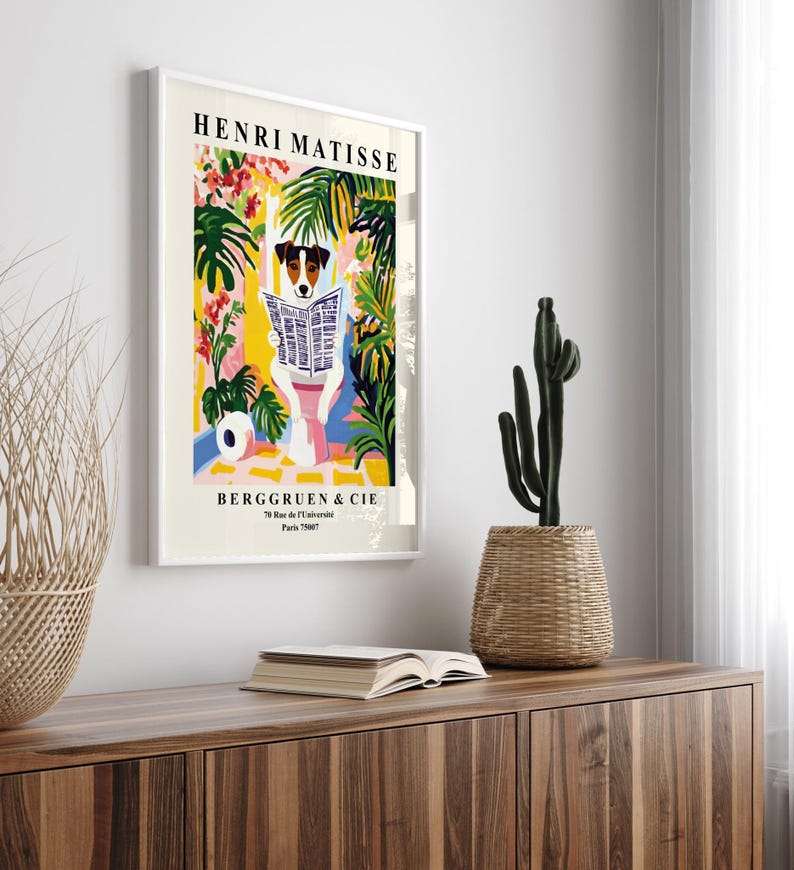 May include: A framed art print featuring a dog reading a newspaper, with the text "HENRI MATISSE" at the top. The artwork has a colorful, floral background. The print is displayed on a wooden cabinet, with a cactus and a woven basket.