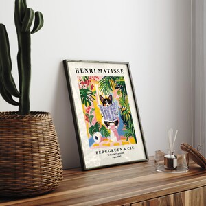 Puede incluir: Impresi&oacute;n art&iacute;stica enmarcada que presenta una ilustraci&oacute;n caprichosa de un corgi leyendo un peri&oacute;dico, sobre un vibrante fondo de follaje tropical. La impresi&oacute;n est&aacute; etiquetada como "HENRI MATISSE" y "BERGGRUEN & CIE". La obra de arte se exhibe sobre una superficie de madera.