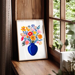 Op de afbeelding: Een ingelijst afdruk van een kleurrijk bloemenboeket in een blauwe vaas. De bloemen zijn geel, oranje, roze en rood. De achtergrond is wit.
