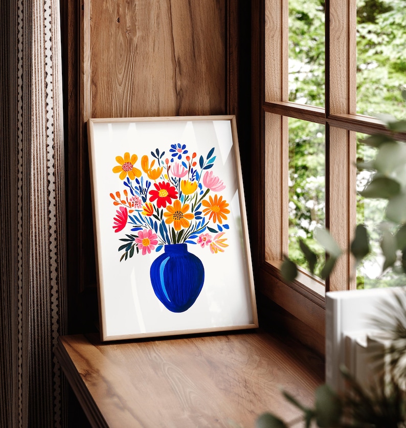 Op de afbeelding: Een ingelijst afdruk van een kleurrijk bloemenboeket in een blauwe vaas. De bloemen zijn geel, oranje, roze en rood. De achtergrond is wit.