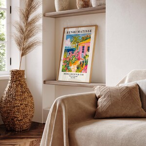May include: A framed poster of a painting by Henri Matisse titled "The Pink House" with a pink house, palm trees, and a blue ocean in the background. The poster is on a shelf with other decorative items.