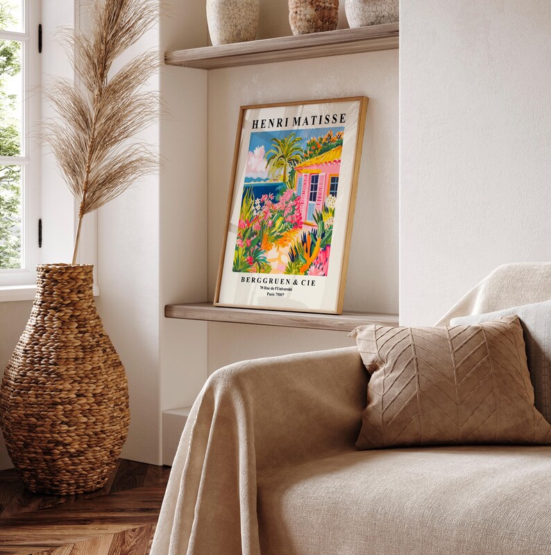 May include: A framed poster of a painting by Henri Matisse titled "The Pink House" with a pink house, palm trees, and a blue ocean in the background. The poster is on a shelf with other decorative items.
