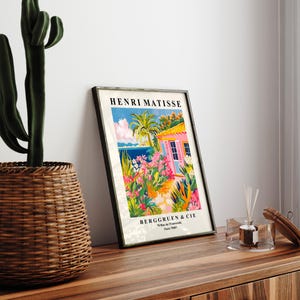 May include: A framed print of a painting by Henri Matisse titled "The Pink House". The painting depicts a pink house with a blue door and a lush garden with palm trees and flowers. The print is framed in black and has the artist's name and the gallery's name and address at the bottom.